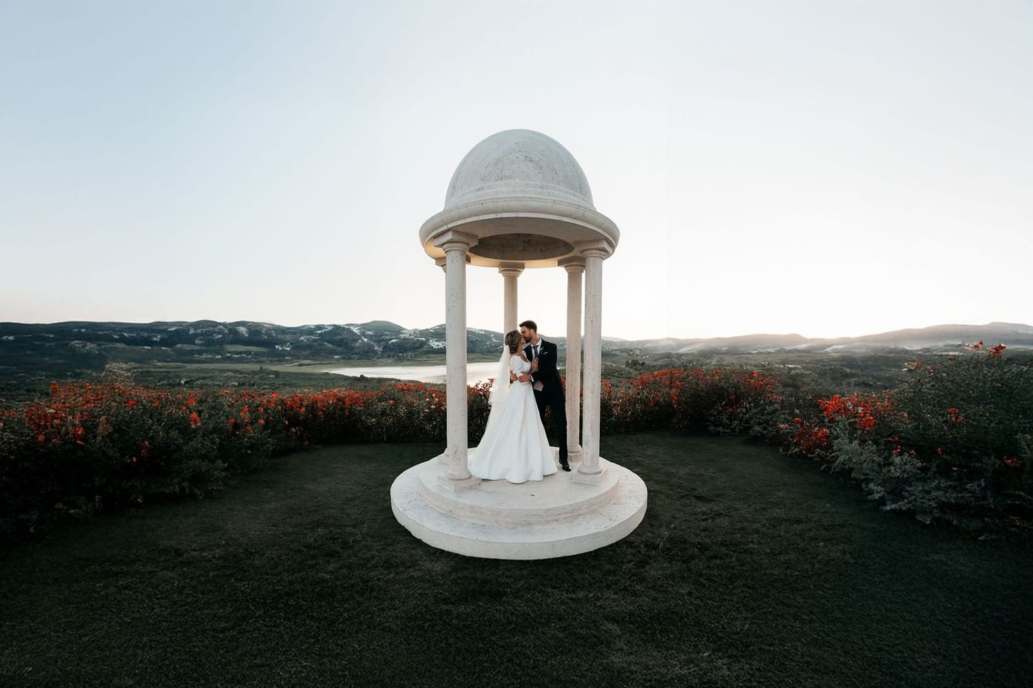 Couple embracing in a white gazebo surrounded by flowers, with mountains and a lake at sunset