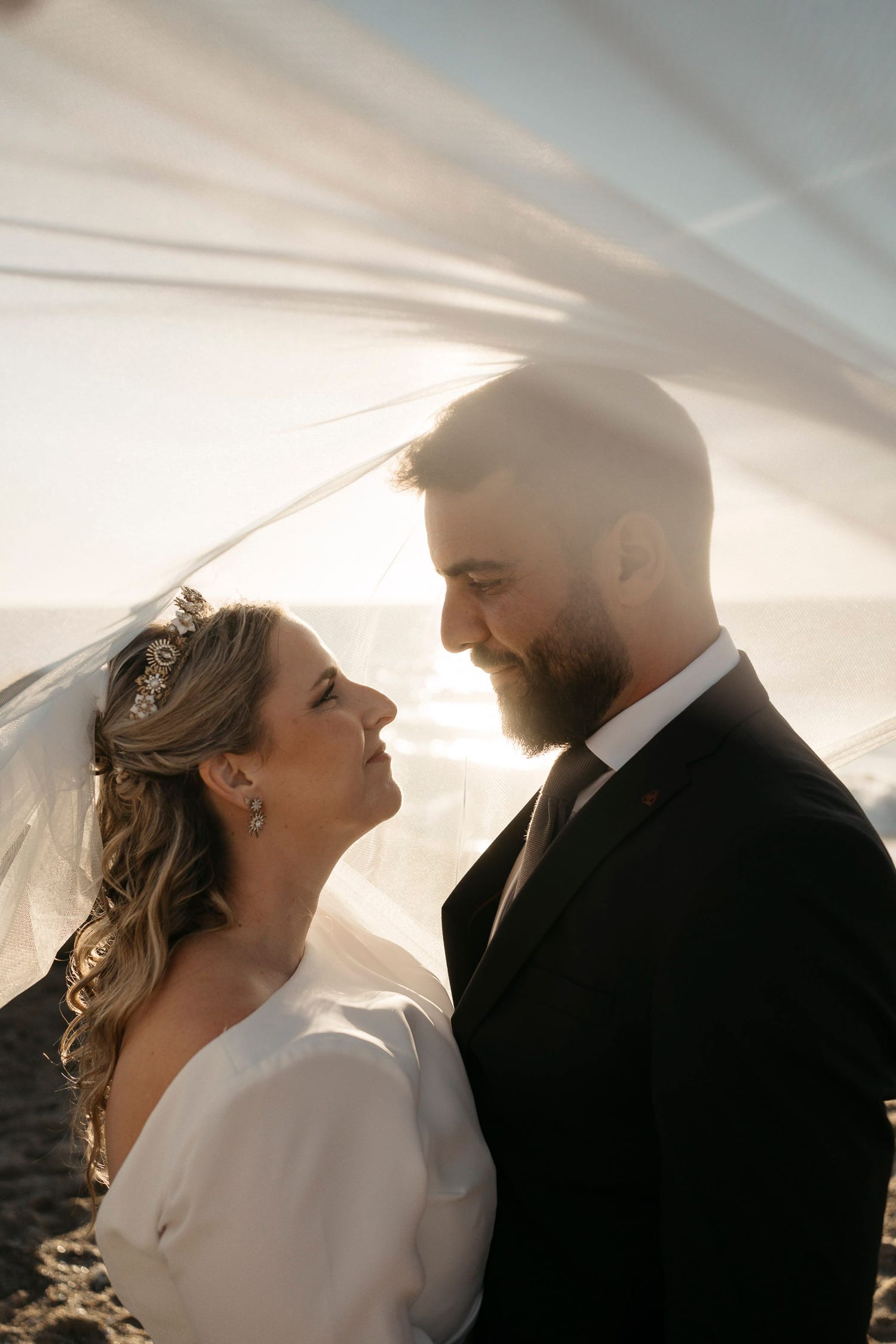 Bride and groom under the veil, backlit in golden light by the sea